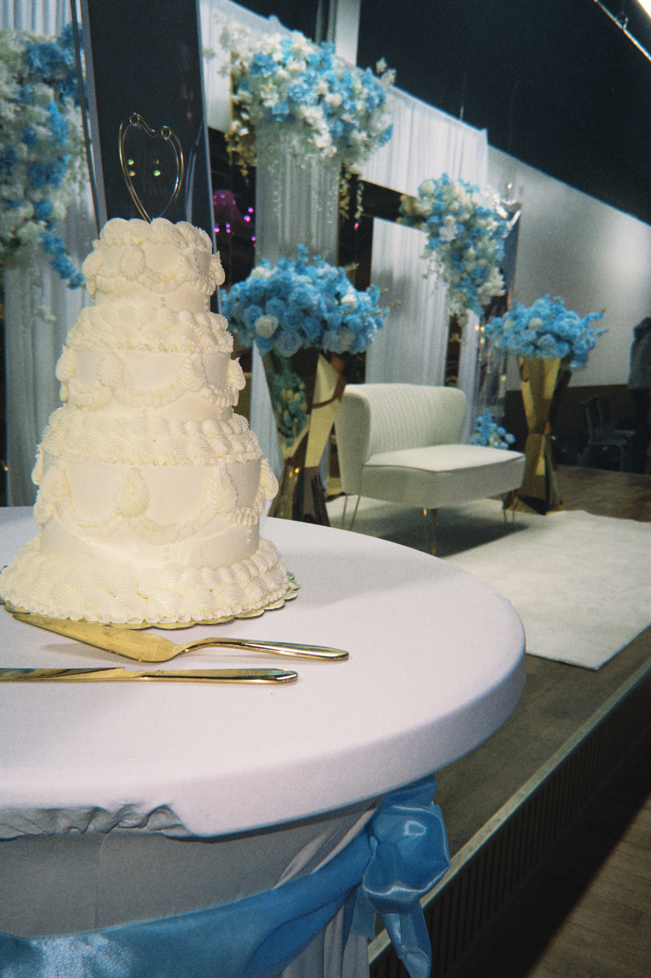 Tiered white wedding cake with piped detail on a draped cake table beside a blue and ivory floral stage.