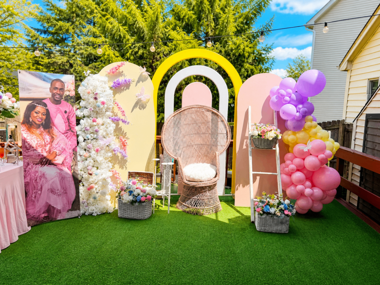 Outdoor celebration setup with layered pastel arches, floral styling, and statement chair.
